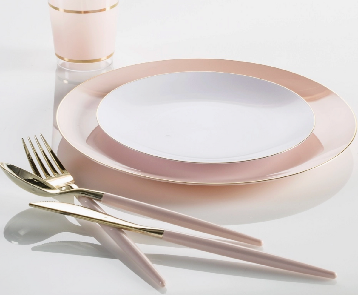 A place setting featuring a large oval charger plate with a wide blush pink rim and gold trim, a smaller white plate in the center, blush pink cutlery with gold detailing, and a matching blush pink cup with a gold band, all arranged on a clean white reflective surface.