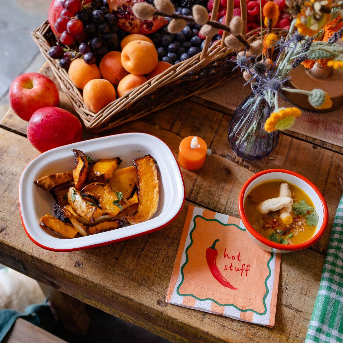 Wooden table with a basket of fruits, a container of roasted vegetables, a bowl of soup, and a card labeled 'hot stuff'.
