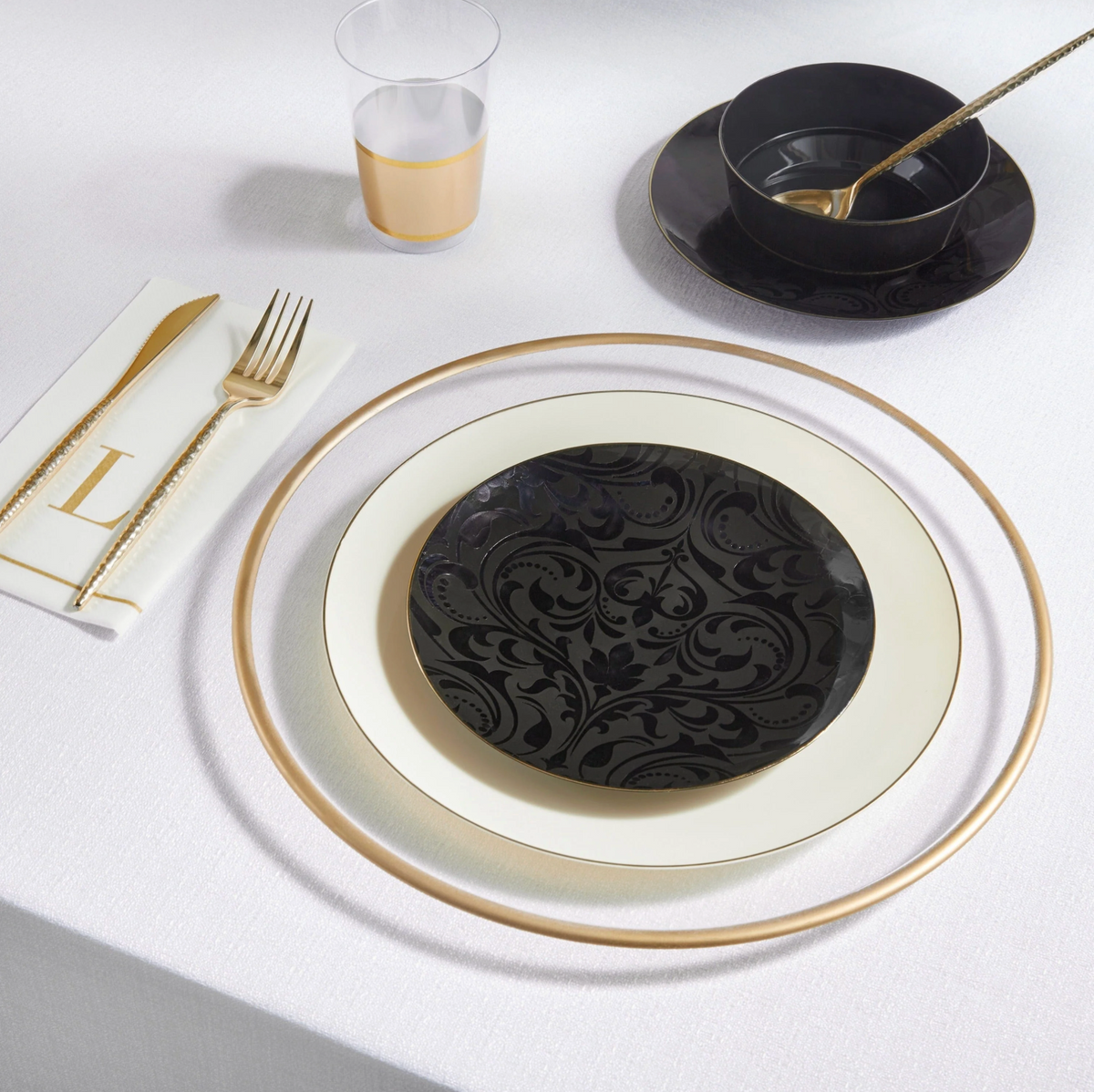 A place setting on a white tablecloth featuring a black oval plate with a gold rim, a cream-colored plate, a gold-rimmed napkin with gold cutlery, a black bowl with a gold spoon, and a clear plastic cup with a gold band.