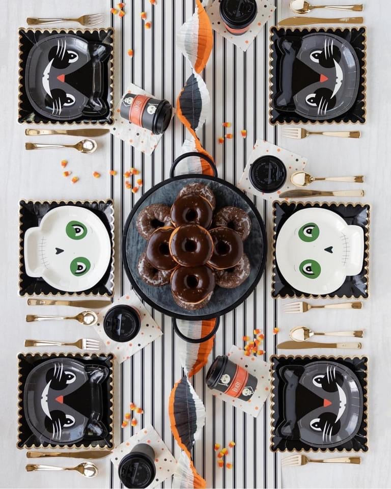 Halloween-themed table setting with white skull and black cat plates, over a striped table runner.