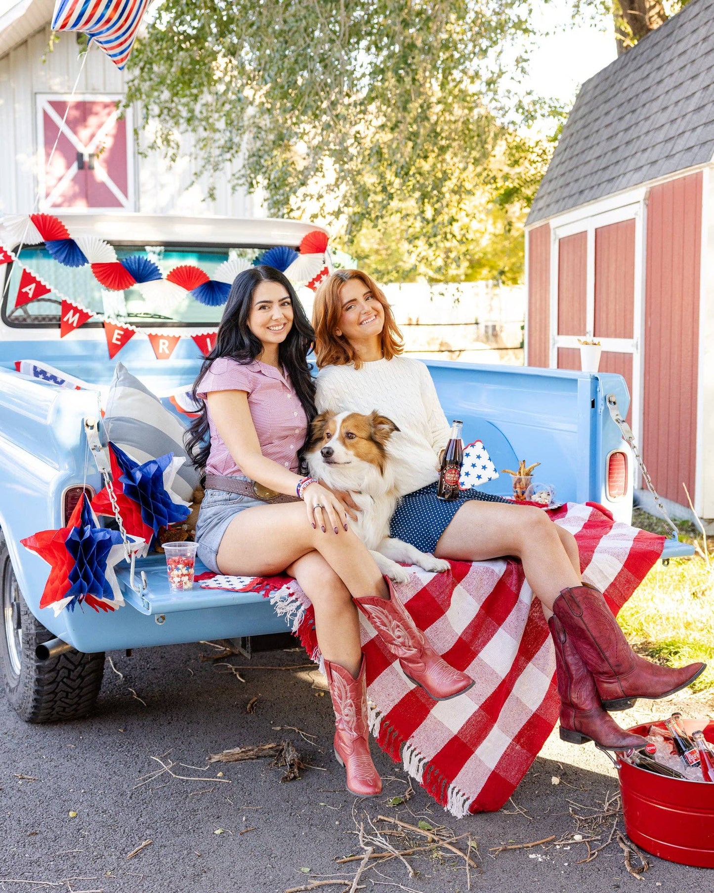 Two women and a dog sitting in the back of a decorated truck with Americana party decorations and a red barn in the background.