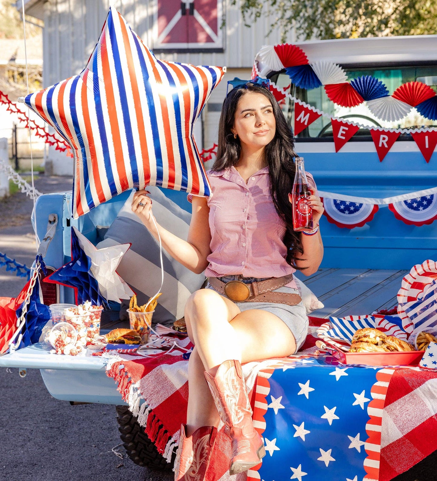 Woman sitting on a truck tailgate with a patriotic theme, holding a star-shaped balloon and a Coca-Cola bottle.