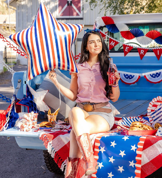 Woman sitting on a truck tailgate with a patriotic theme, holding a star-shaped balloon and a Coca-Cola bottle.