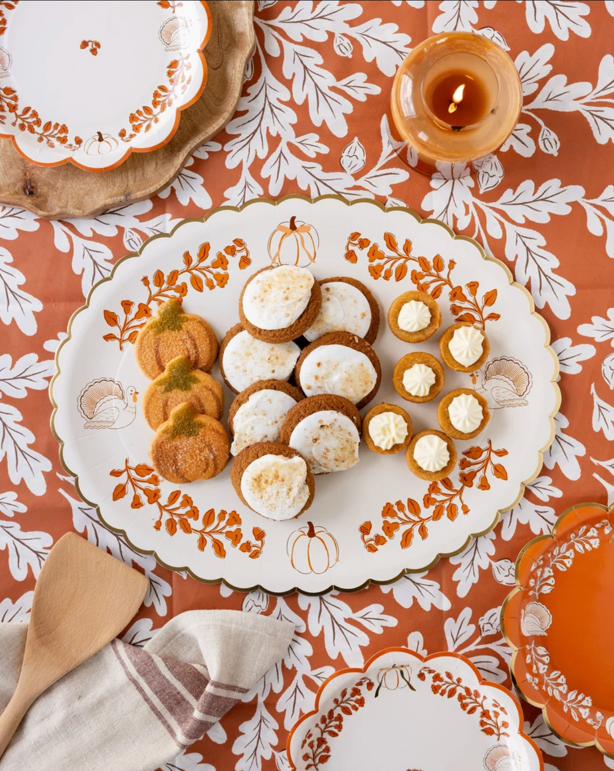 A styled overhead flat-lay of a white scalloped serving platter with orange and gold fall leaf and pumpkin motifs, filled with cookies and cream canapes, set on an orange tablecloth with matching plates and a lit candle.
