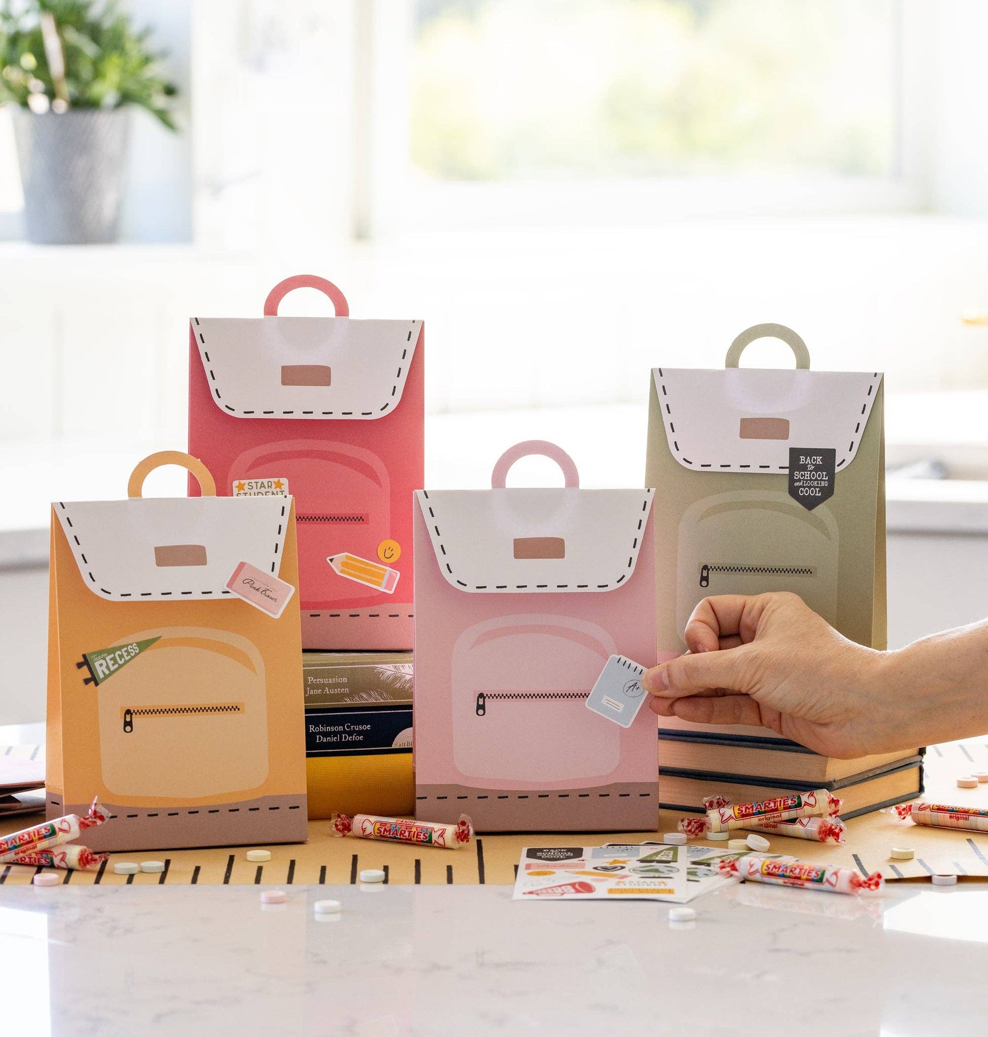 A set of four Back to School themed treat bags in pink, green, yellow, and orange colors, with a decorative sticker sheet, displayed on a table.