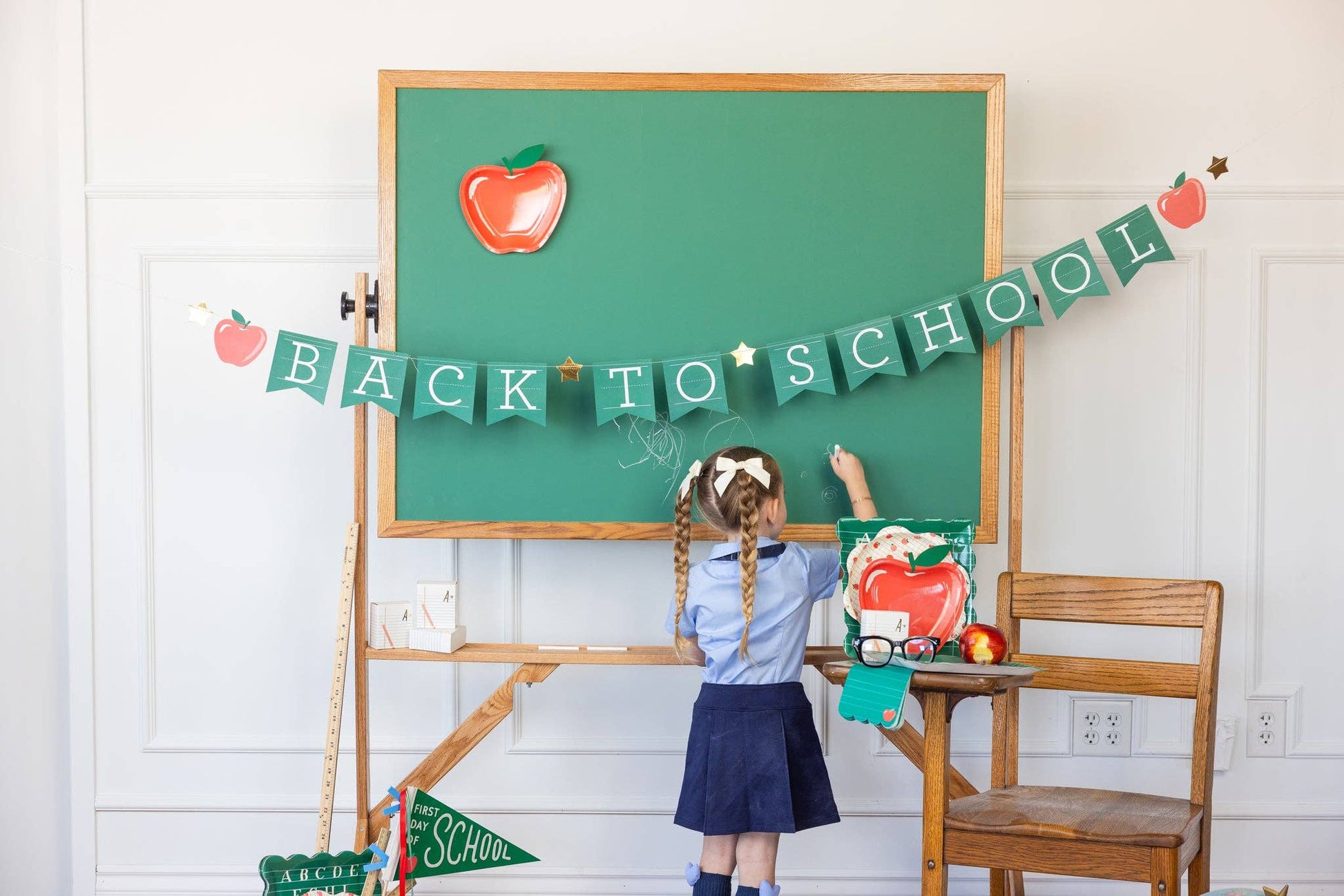 Child in school uniform standing in front of a green chalkboard with 'Back to School' decorations.