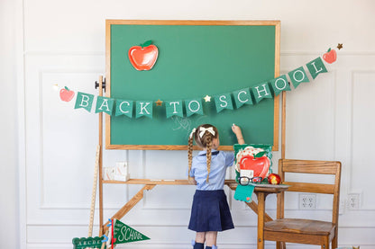 Child in school uniform standing in front of a green chalkboard with 'Back to School' decorations.