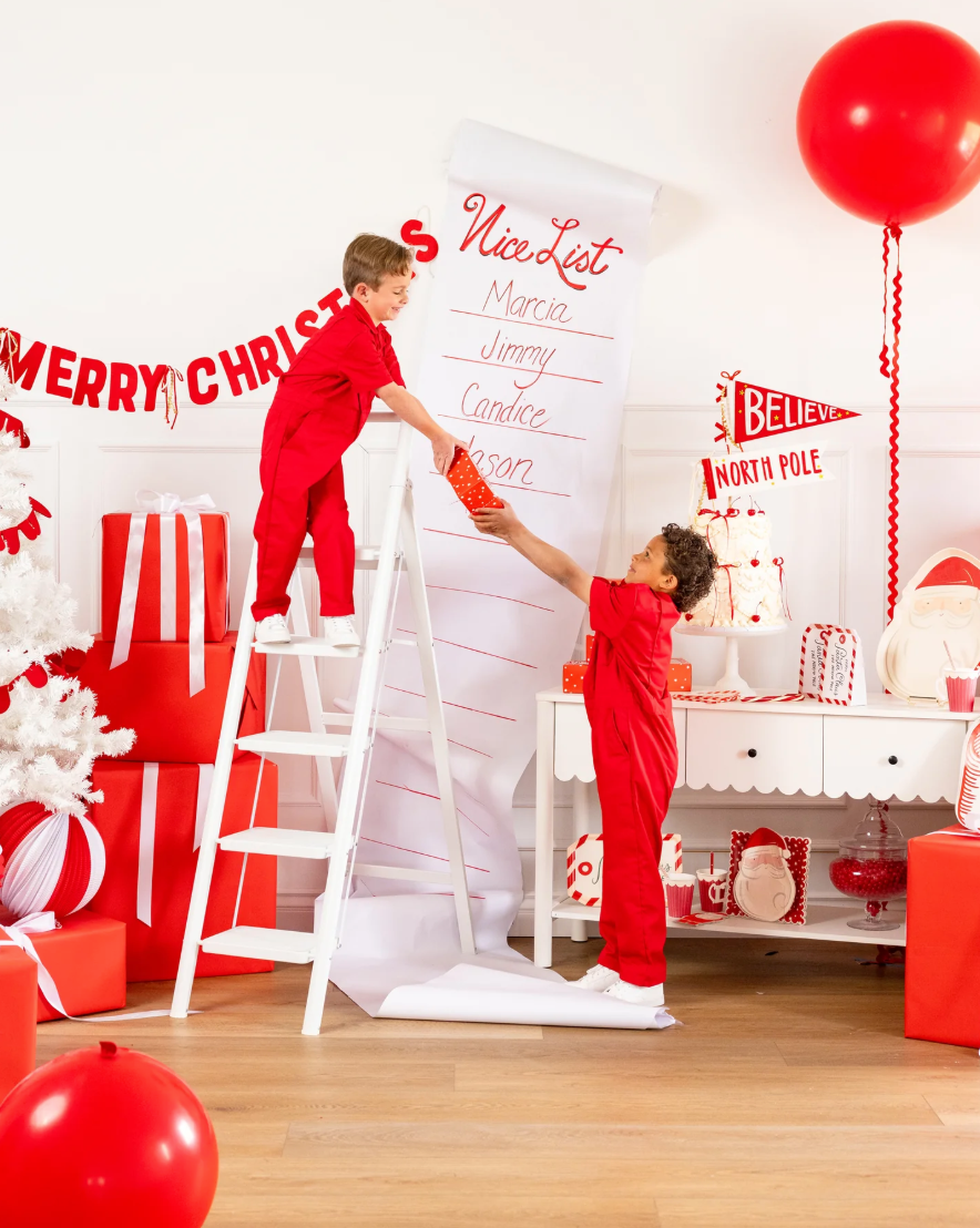 Two children in red outfits decorating a Christmas-themed room with balloons and presents.