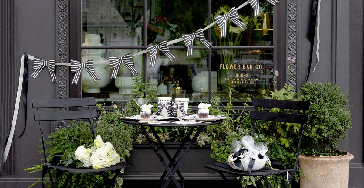 Outdoor seating area with a small table, chairs, and decorative elements in front of a store window with a paper bow banner.