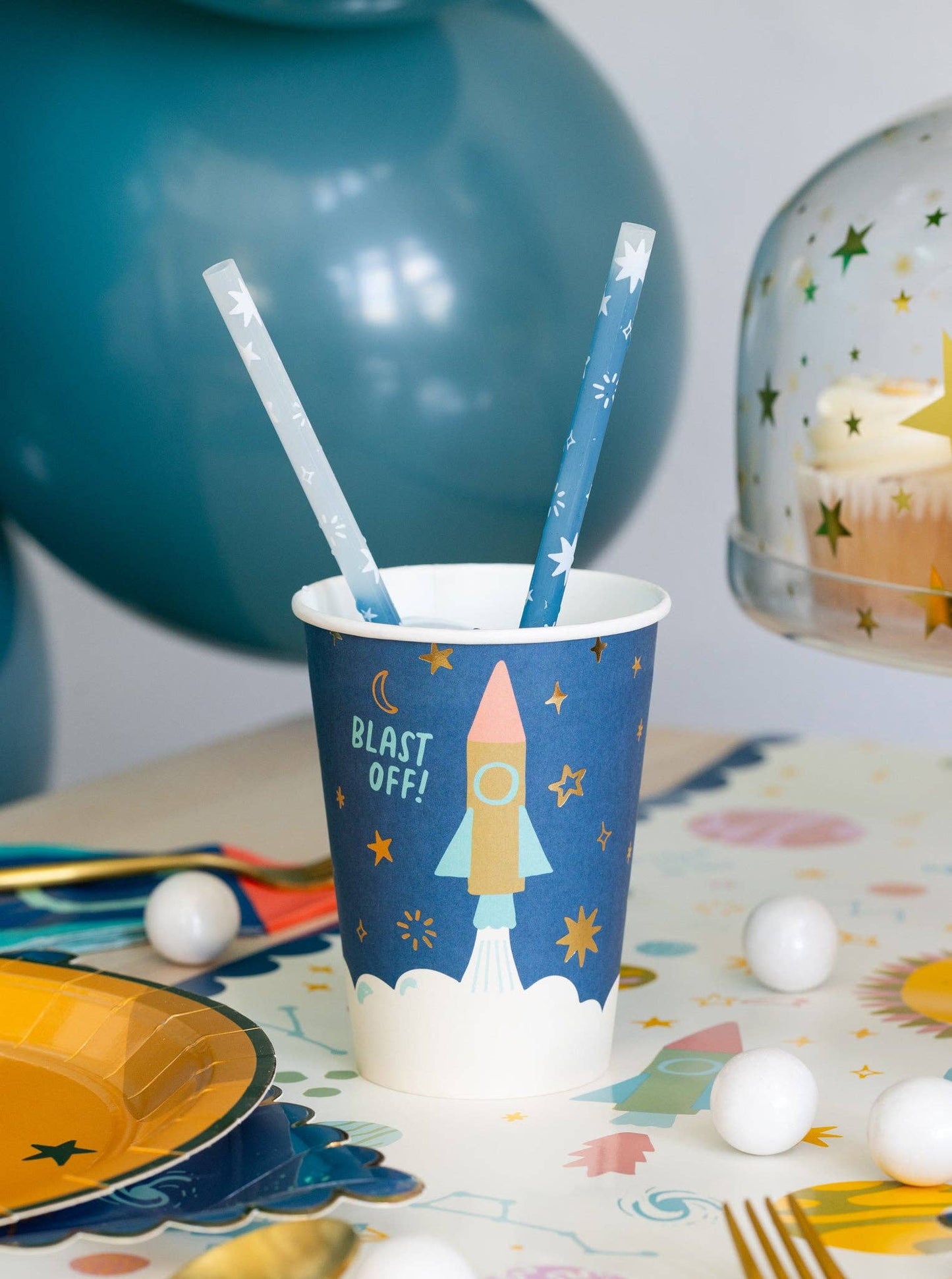 Party cup with rocket design and straws on a table with balloons and cake in the background