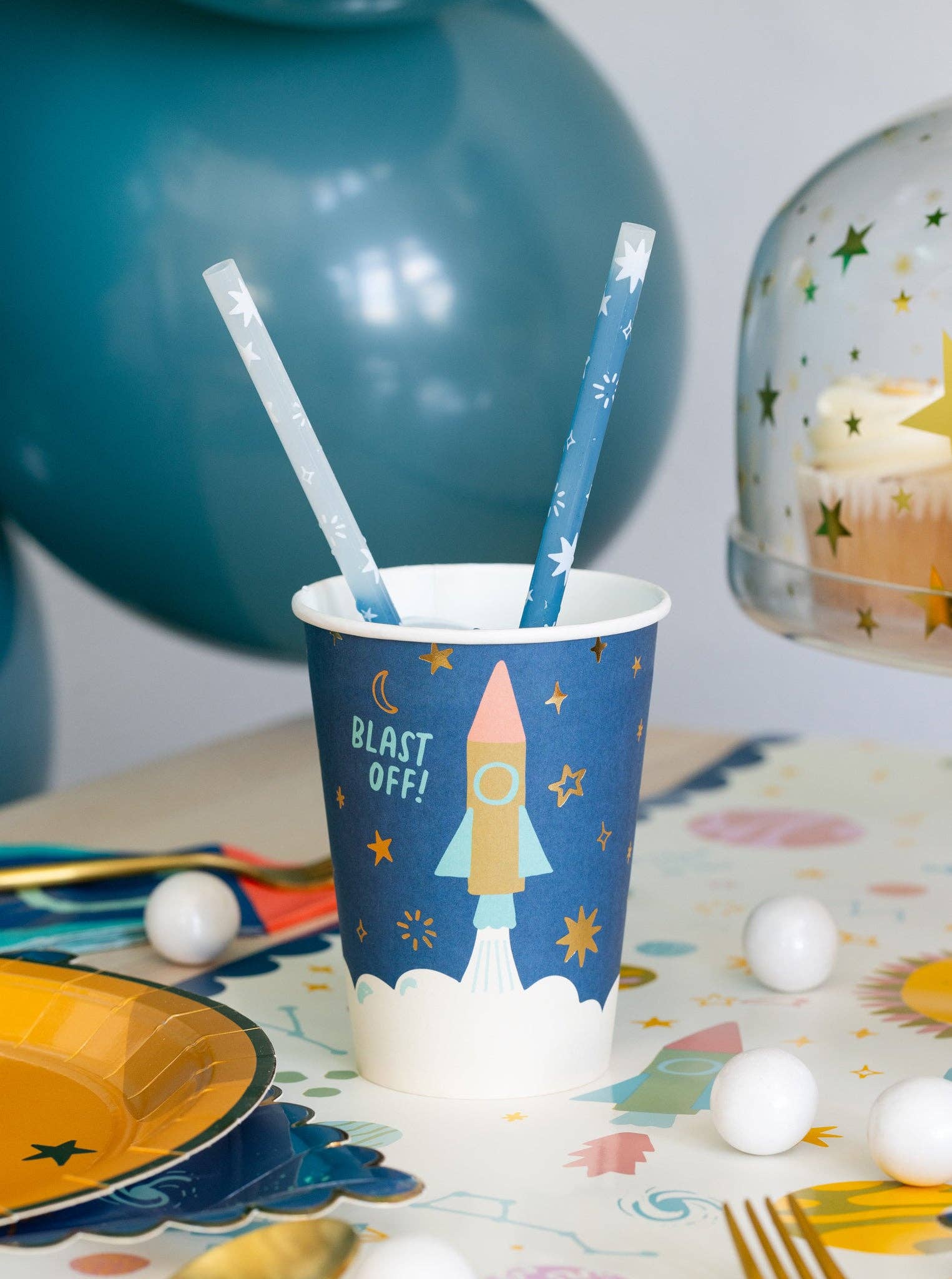 Party cup with rocket design and straws on a table with balloons and cake in the background