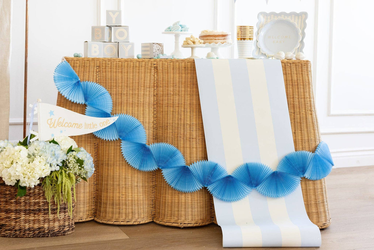 Decorative blue tissue paper accordion-style garland on a brown wicker table with flowers and boy baby shower decor items.
