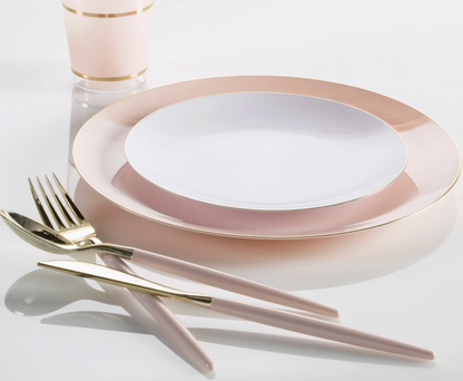 A place setting featuring a large oval charger plate with a wide blush pink rim and gold trim, a smaller white plate in the center, blush pink cutlery with gold detailing, and a matching blush pink cup with a gold band, all arranged on a clean white reflective surface.