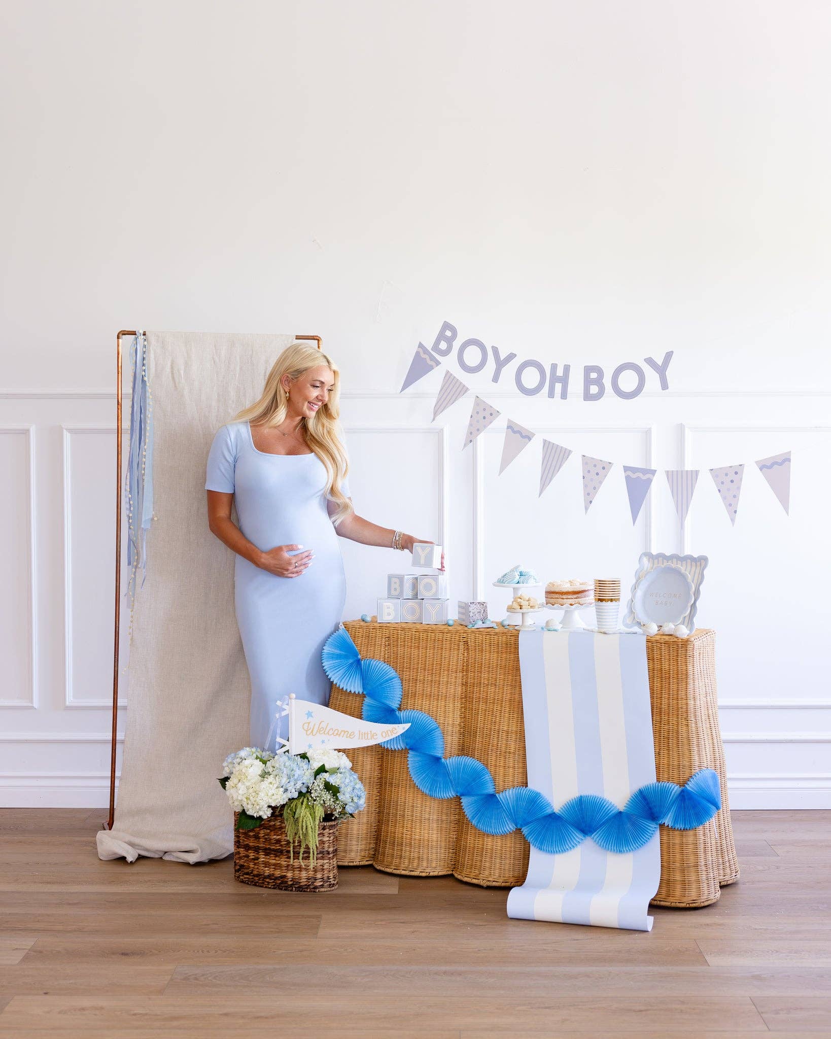 Pregnant woman in a light blue dress standing next to a decorated table with 'BOY OH BOY' banner for a baby shower.