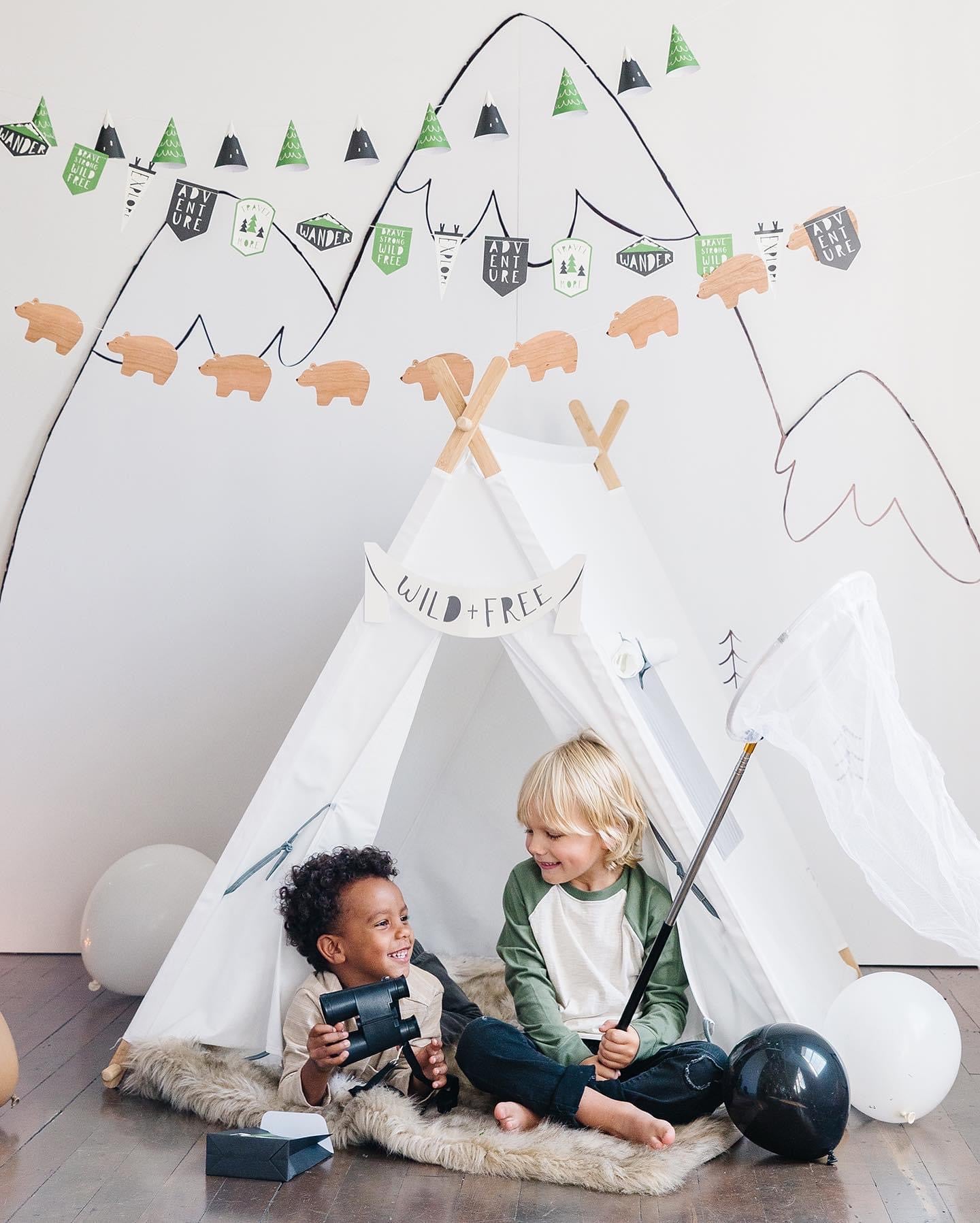 Two children playing inside a white tent with camping adventure-themed party decorations on a light gray wall.