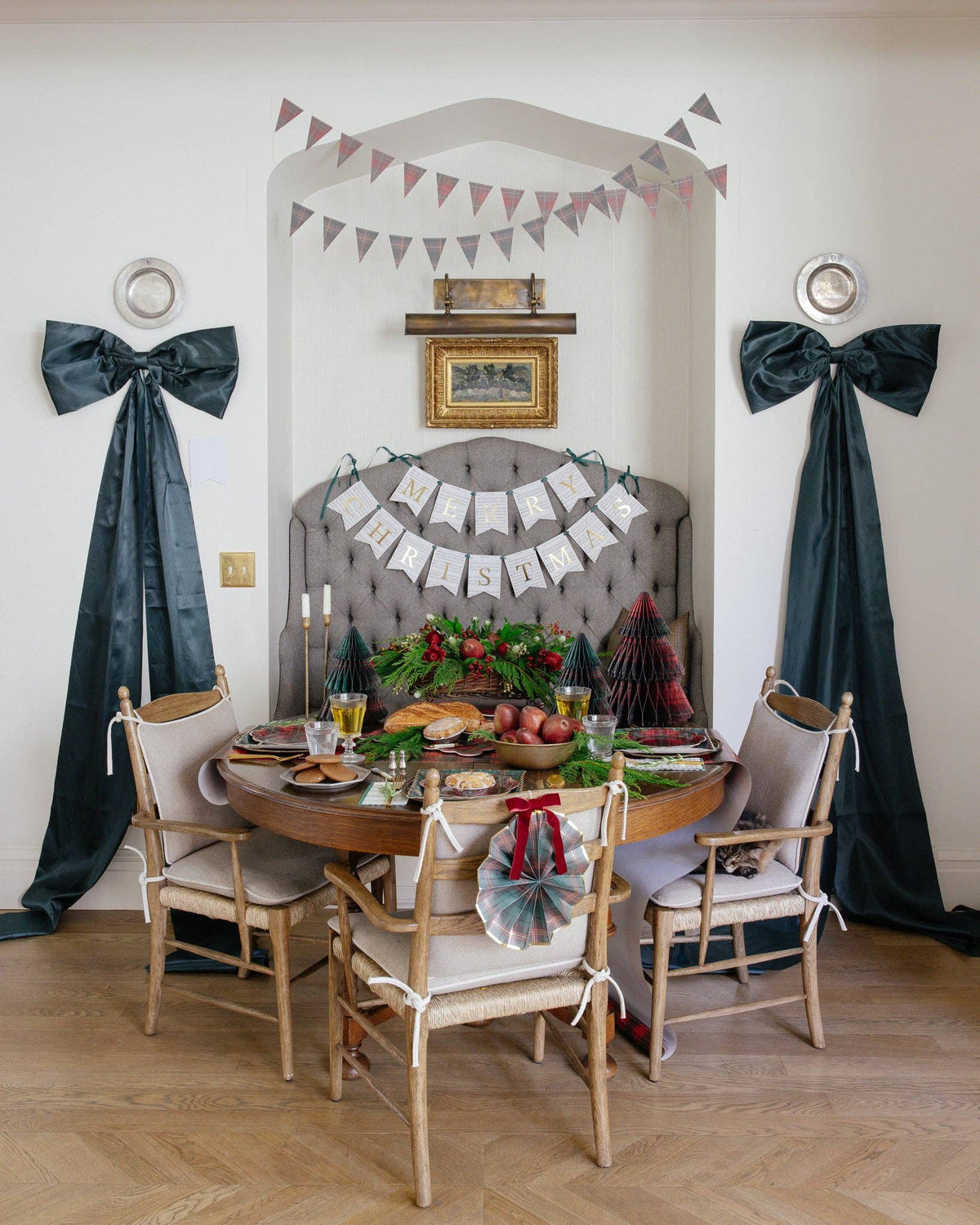 A styled dining room scene with a round wooden table set for a meal, four light wood chairs with cream upholstery, dark teal velvet bows hanging on both sides, and a gray tufted headboard backdrop with bunting and holiday decorations.