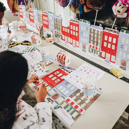 A child sits at a table crafting with colorful paper city buildings, small toy cars, and printed activity sheets in a bright indoor Christmas party setting.