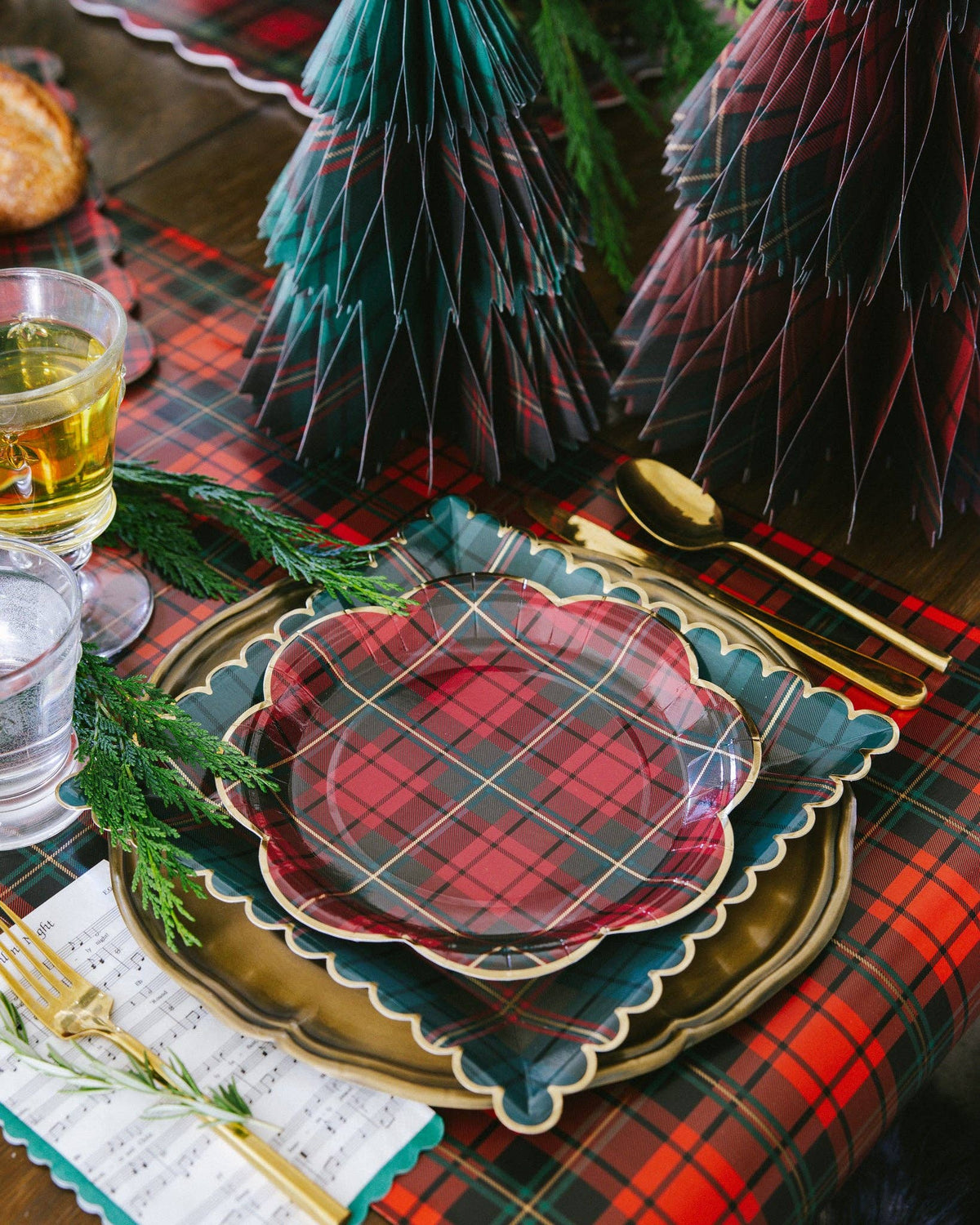 A styled holiday table setting with red and teal plaid plates, gold cutlery, green pine branches, and tartan tablecloth in a warm, festive setting.