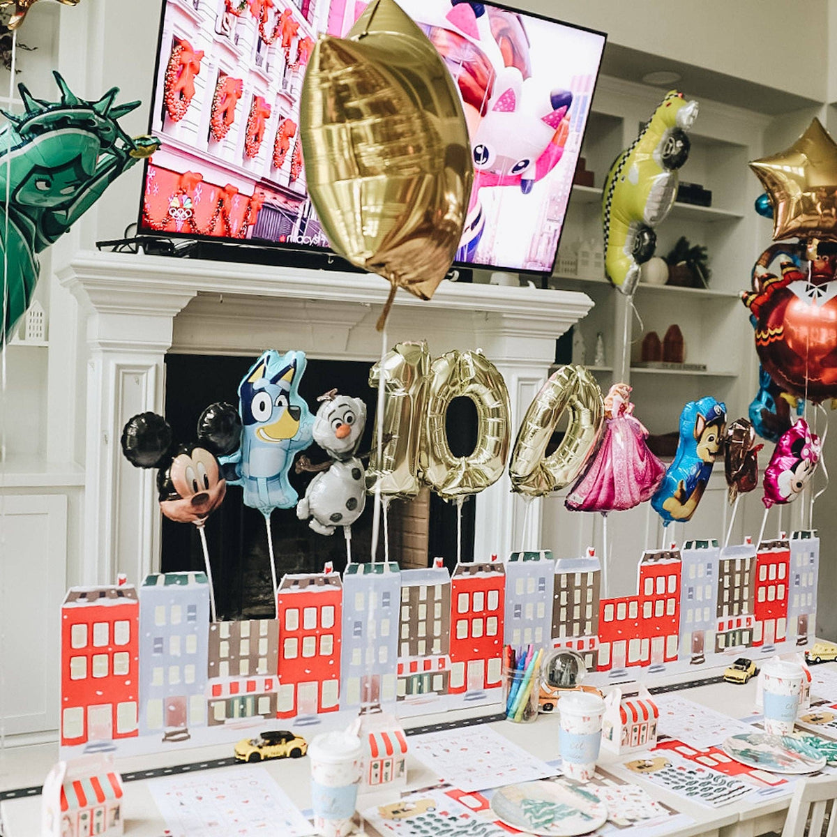 A decorated party table in a living room with a white fireplace mantel and TV in the background. The table is set with colorful character balloons, city building paper cups, plates, napkins, and small toy cars, creating a festive Holiday atmosphere.