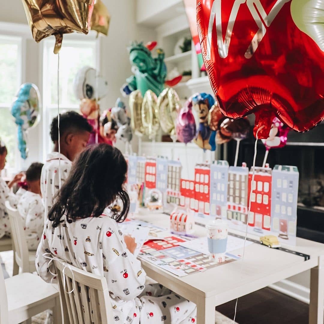 Children in white patterned pajamas sit at a decorated table with colorful balloons, toy houses, and party supplies in a bright indoor room.