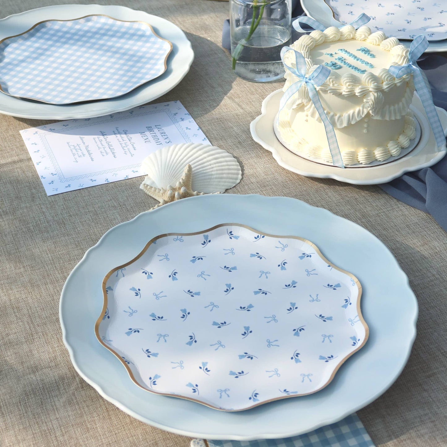 Decorative table setting with a blue plate featuring white floral patterns, a cake, and shell decorations on a textured surface.