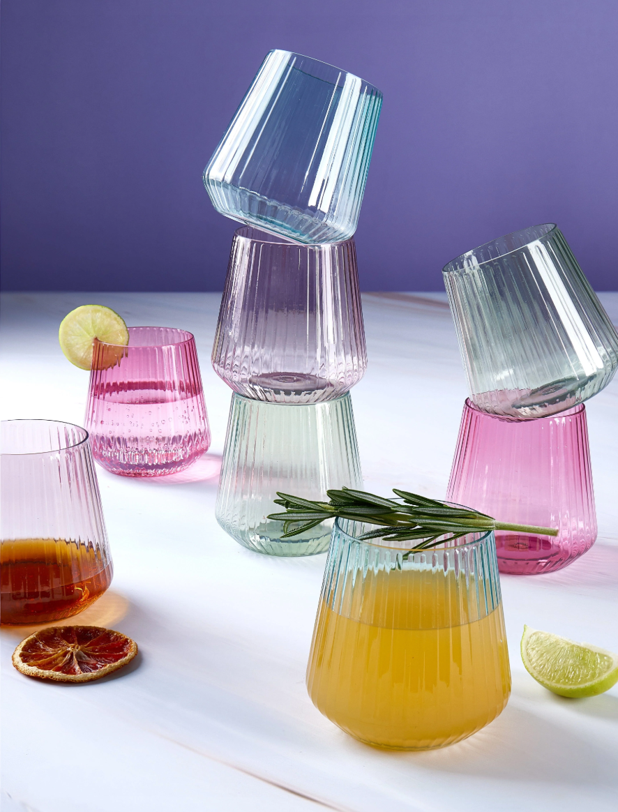 Colorful glass stemless cups with drinks and garnishes on a white surface against a purple background