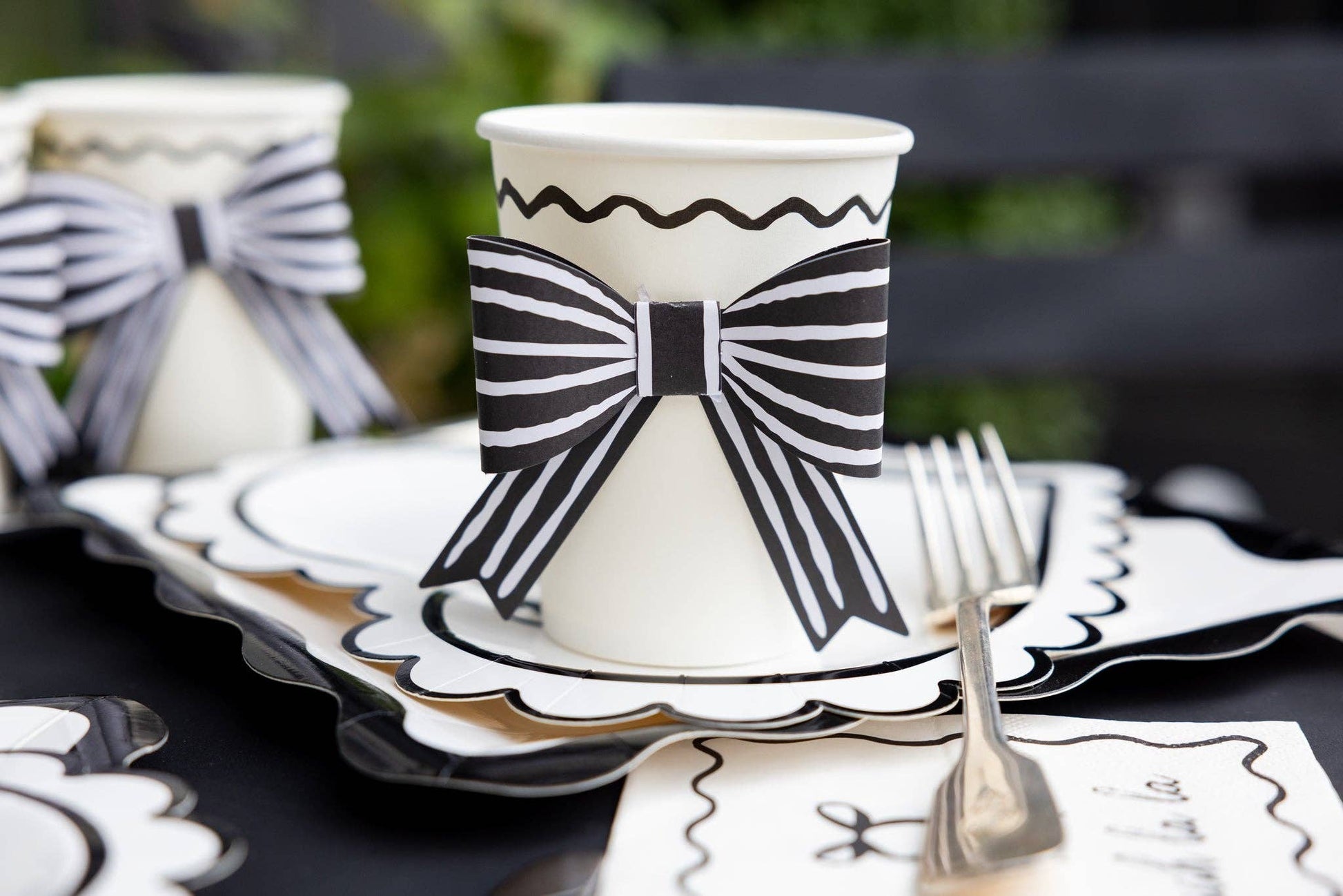 Cream colored paper cup with 3D black bow decoration on a table setting with paper plates and napkins.