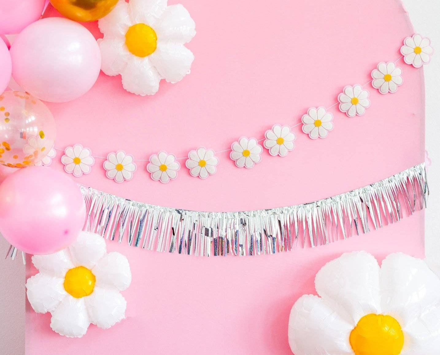 Retro party decoration setup featuring a silver tinsel fringe banner, a string of white daisy flower garlands with yellow centers, and pink balloons, including one clear balloon with gold confetti.
