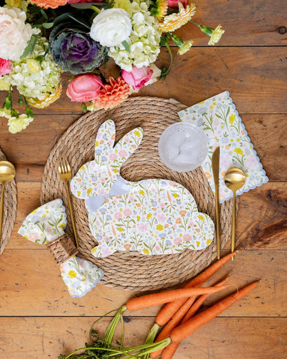 Easter-themed place setting with floral bunny plates and matching napkins, carrots, and flowers on a wooden table.