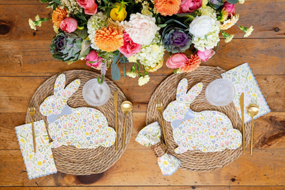 Two woven placemats with floral bunny paper plates and napkins on a wooden table, accompanied by a bouquet of flowers.