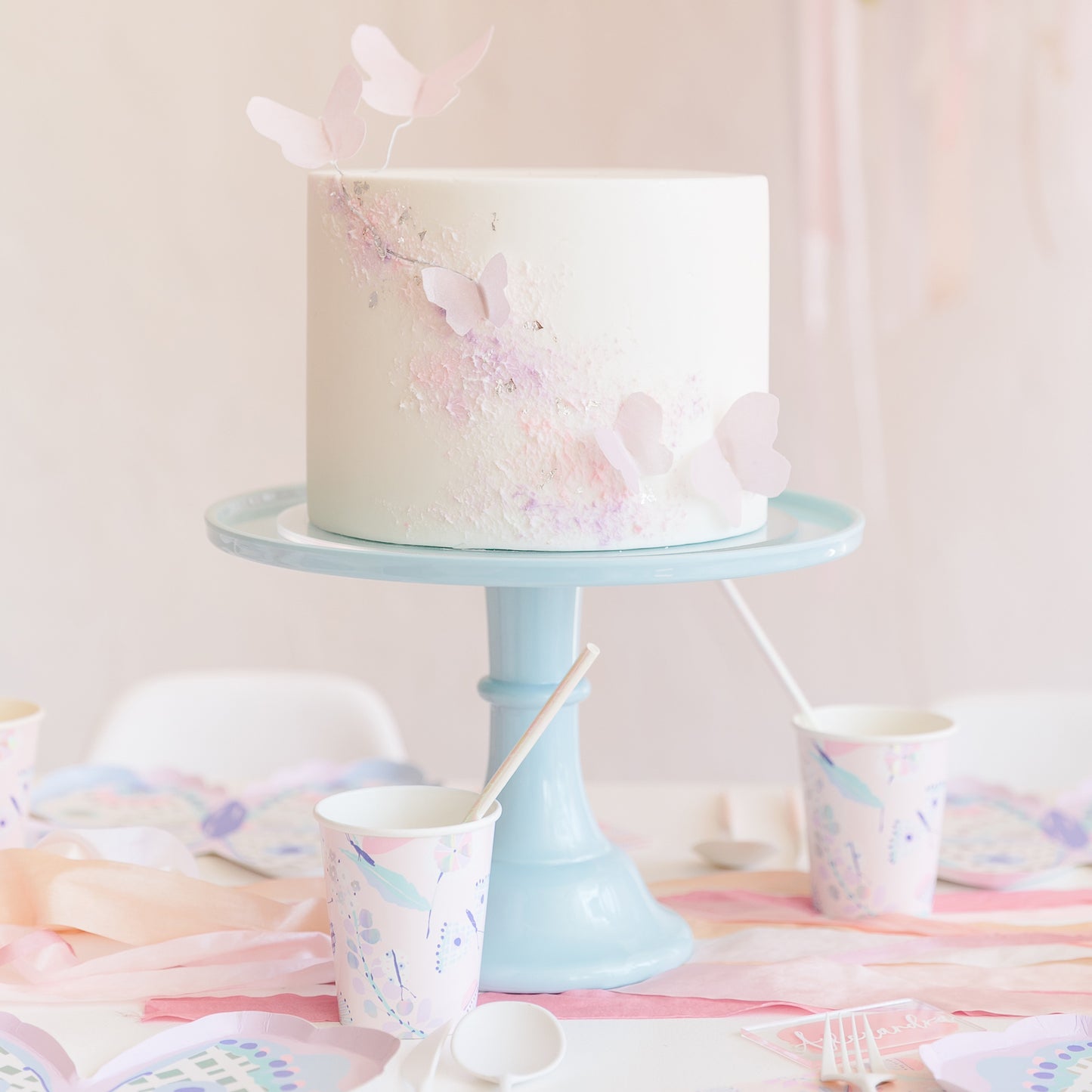 A white frosted birthday cake decorated with pastel pink and lavender butterfly and glitter accents, displayed on a light blue cake stand on a decorated party table with matching paper cups and plates.
