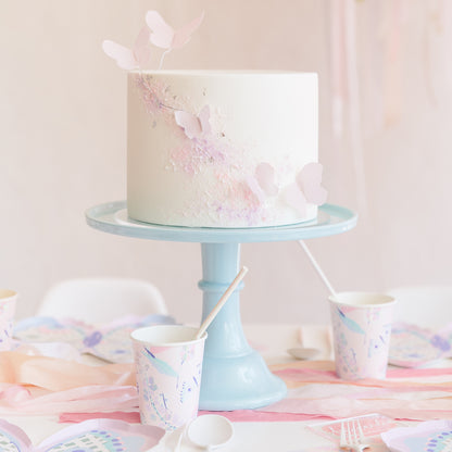 A white frosted birthday cake decorated with pastel pink and lavender butterfly and glitter accents, displayed on a light blue cake stand on a decorated party table with matching paper cups and plates.