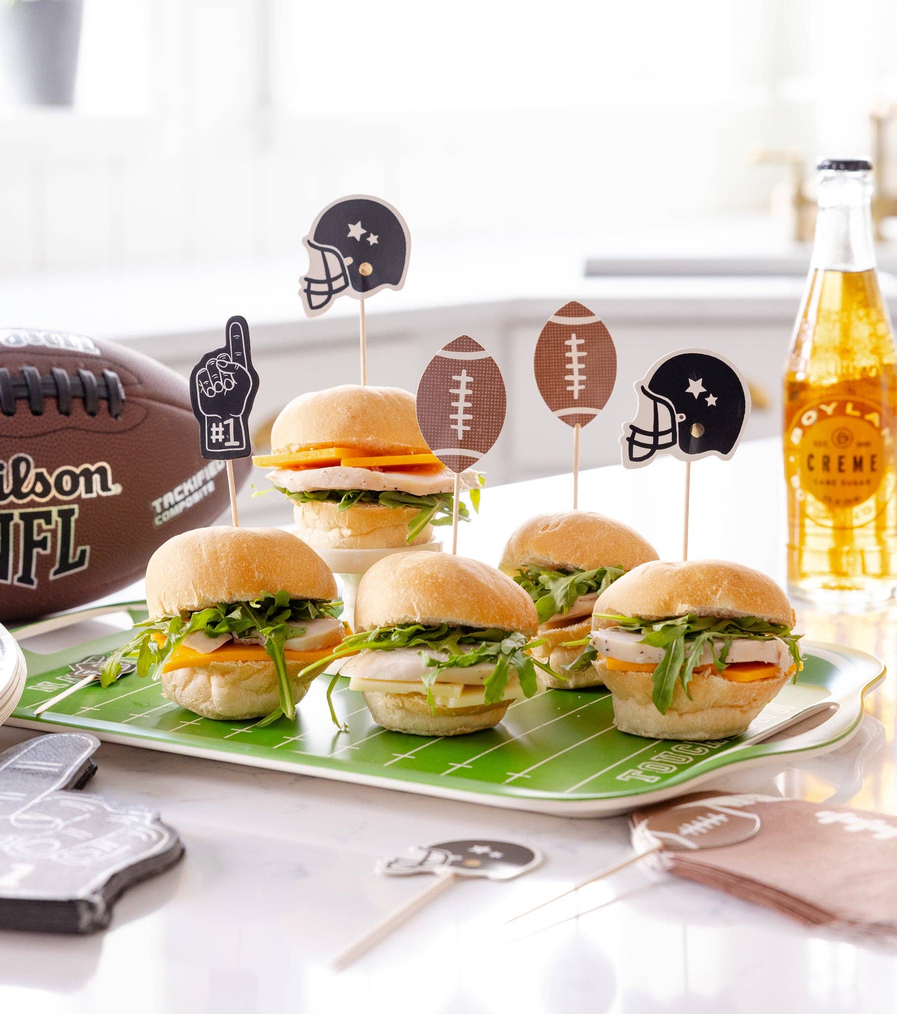 Mini burgers on a platter with football-themed picks, a bottle of cream soda, and a football in the background.