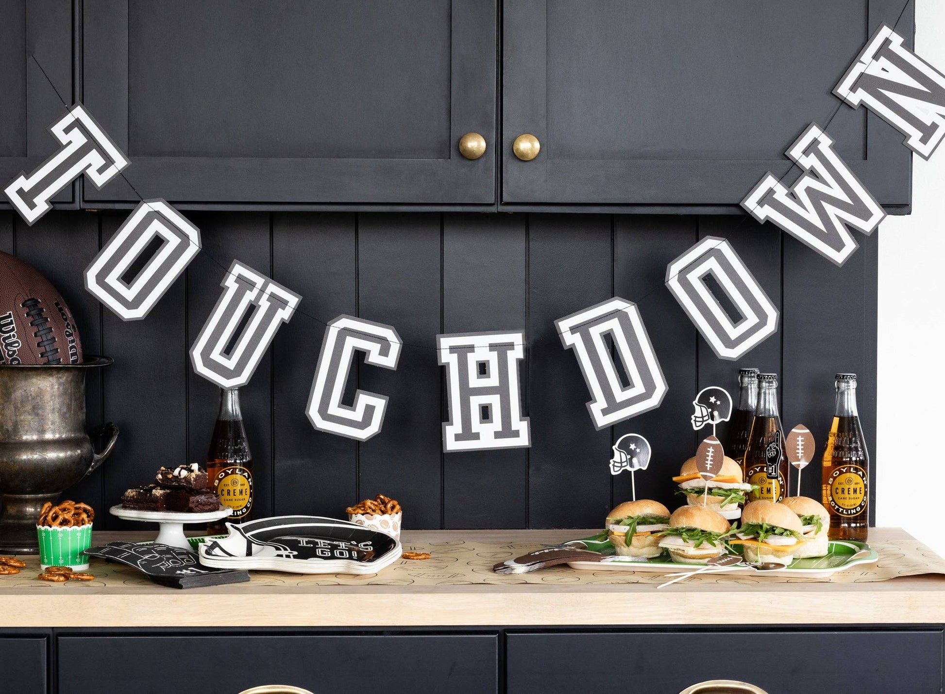 Football party setup with food and drinks on a table against a black cabinetry with a 'TOUCHDOWN' banner.