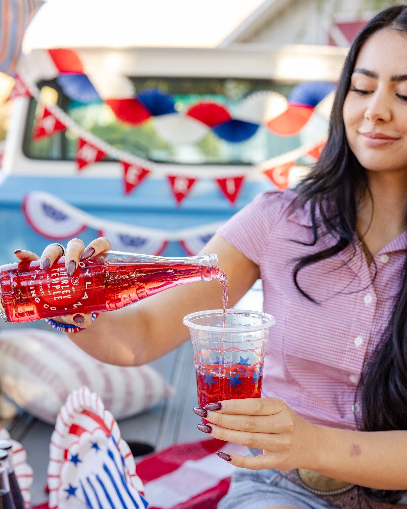 Woman pouring red liquid from a bottle into a cup at a festive outdoor event with patriotic decor.