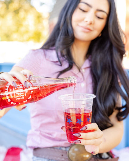 Woman pouring red juice from a bottle into an Americana-themed plastic cup outdoors