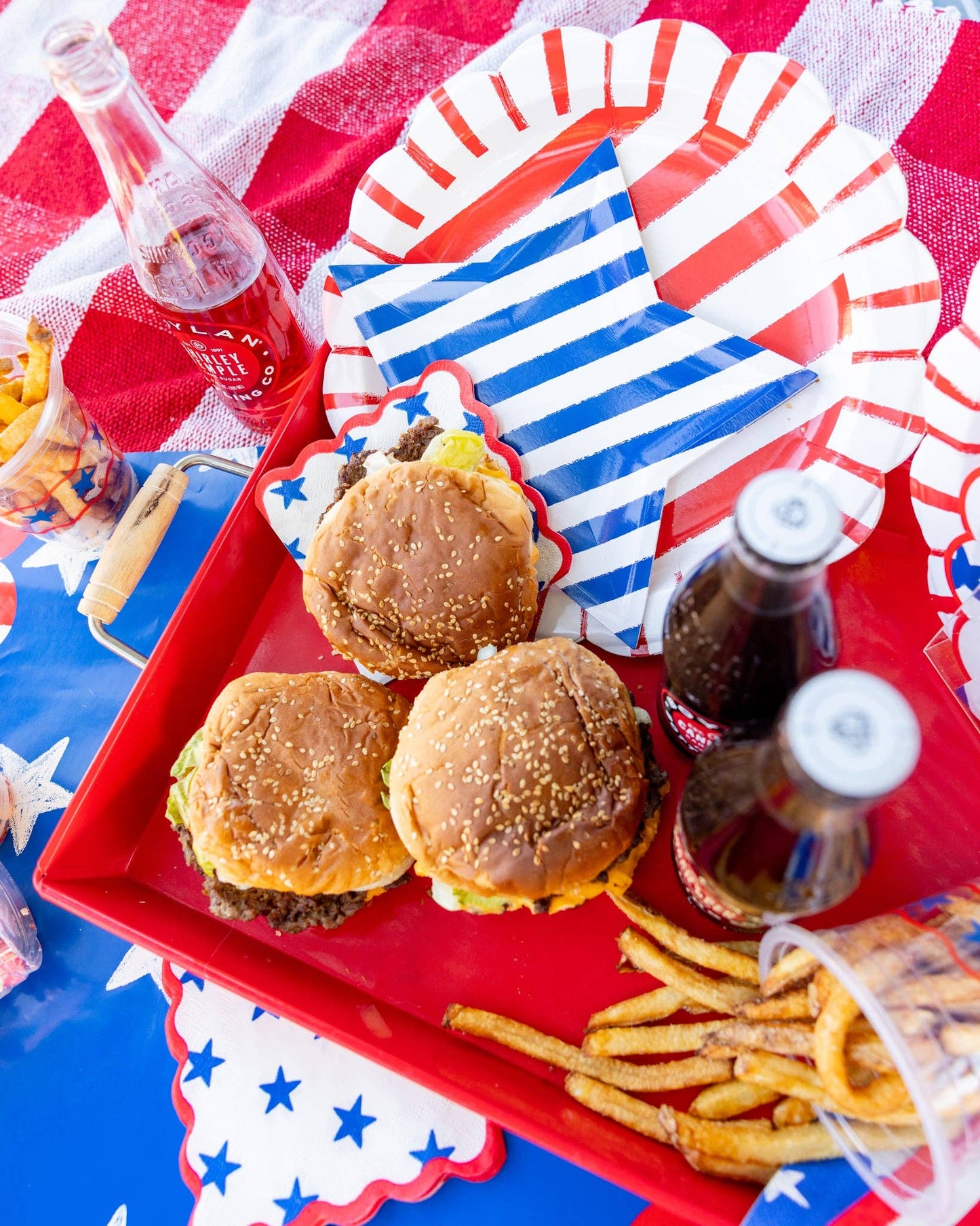 Hamburgers and fries on a red tray with soda bottles, all on a tablecloth with red, white, and blue patterns.