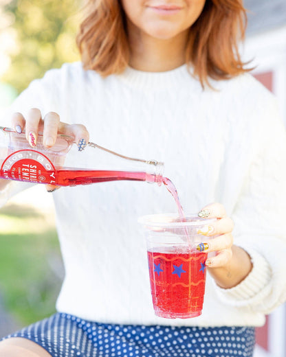 Person pouring a red drink from a glass bottle into a clear plastic cup.
