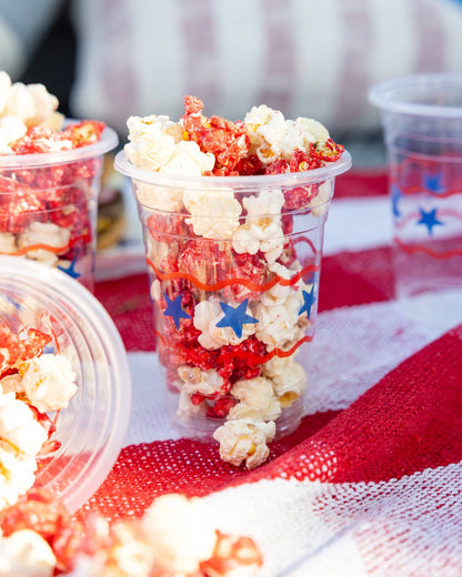 Clear plastic cups filled with red, white, and blue popcorn on a red and white checkered tablecloth.