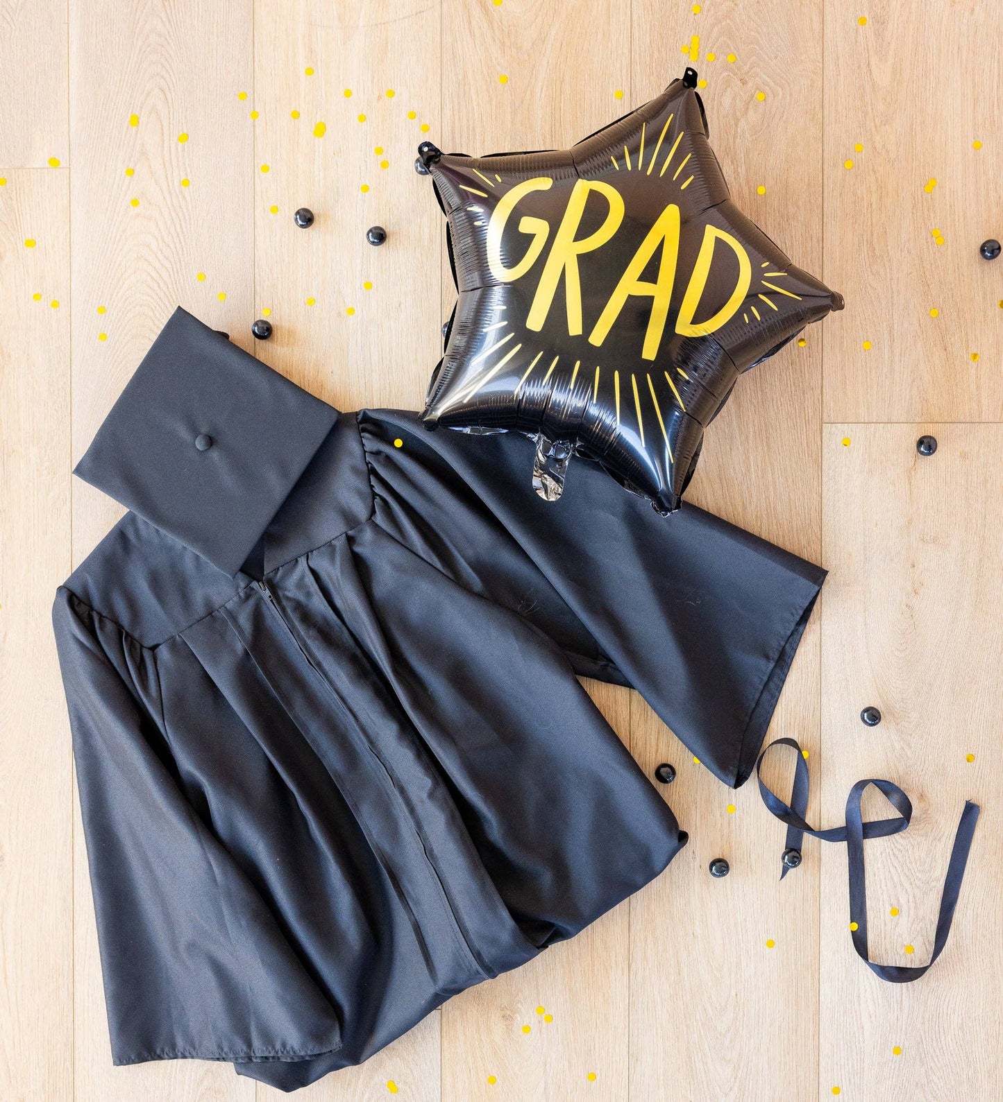 Graduation cap, gown, and 'GRAD' balloon on a wooden surface with confetti.