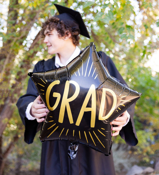 Graduate holding a black star-shaped balloon, with gold text that spells 'GRAD', outdoors with trees in the background.