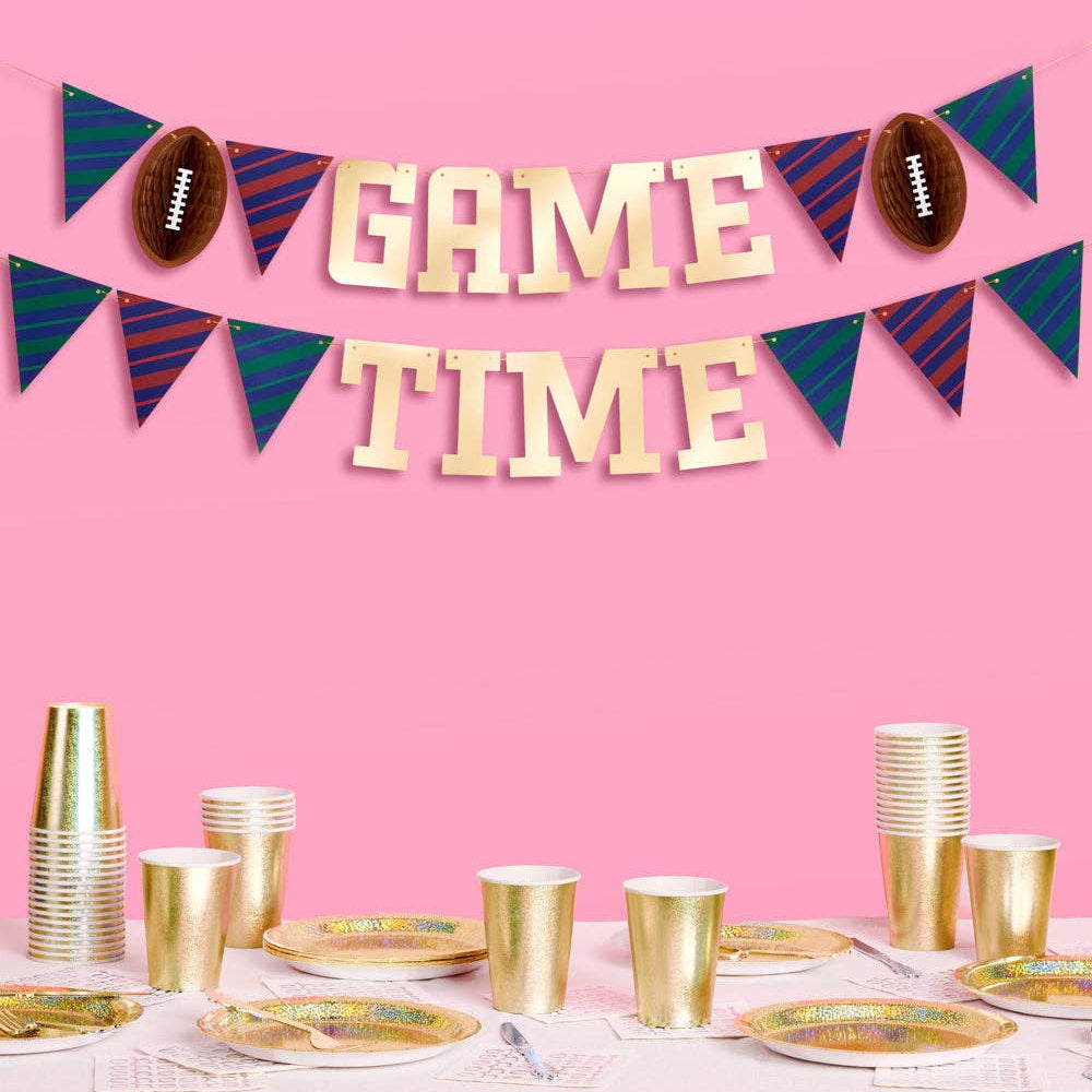 Decorative 'Game Time' banner with footballs and pennants above a table set with gold cups and plates on a pink background.