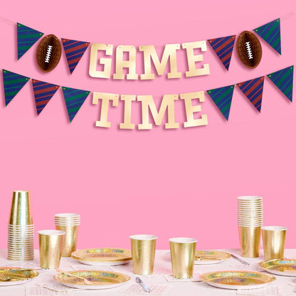 Decorative 'Game Time' banner with footballs and pennants above a table set with gold cups and plates on a pink background.