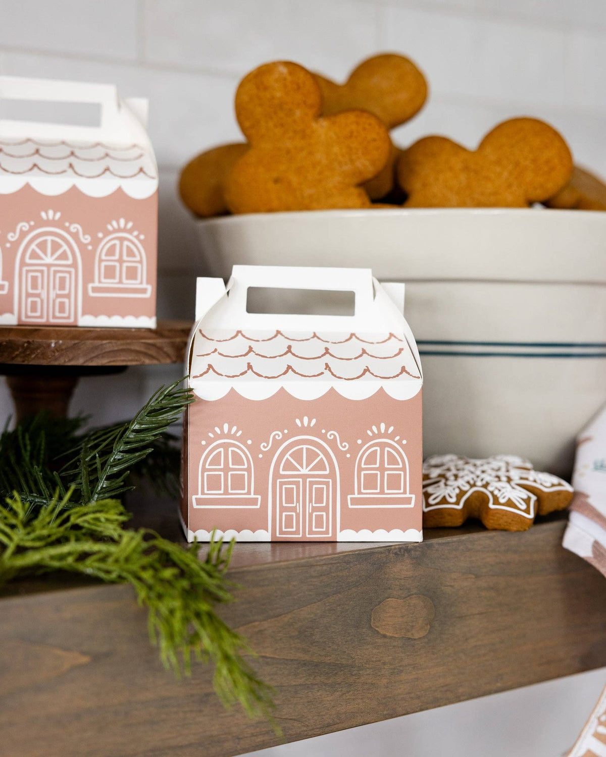 Decorative gift box with gingerbread house design on a wooden surface with cookies in the background.