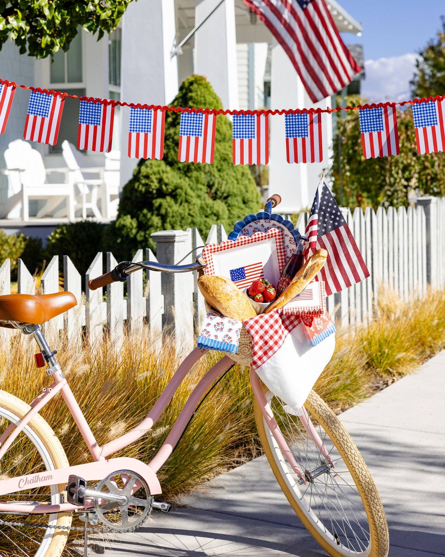 Pink bicycle with a basket of fruit and American flag banner on a white picket fence background