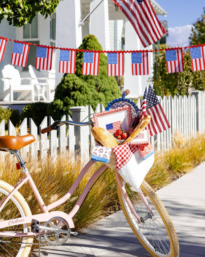 Pink bicycle with a basket of fruit and American flag banner on a white picket fence background