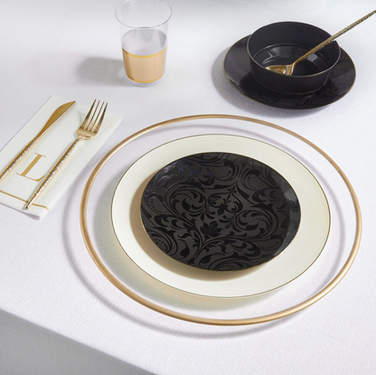 A place setting on a white tablecloth featuring a black oval plate with a gold rim, a cream-colored plate, a gold-rimmed napkin with gold cutlery, a black bowl with a gold spoon, and a clear plastic cup with a gold band.