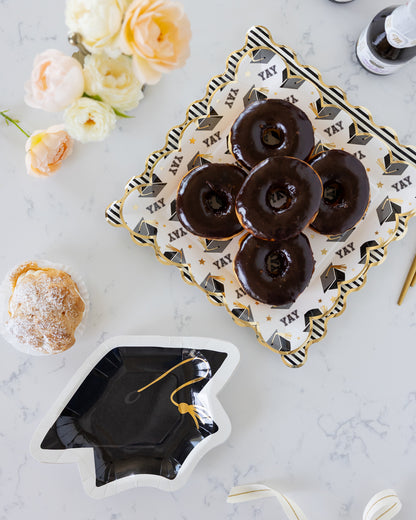 Chocolate donuts on a decorative graduation-themed plate with flowers and a black graduation cap plate nearby.
