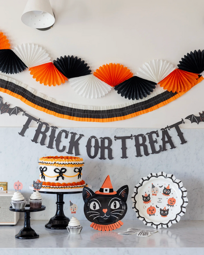 Halloween party decorations displayed on a tabletop, including a black, orange, and white paper fan garland, a black 'Trick or Treat' banner, a black cat face plate, a black cat plate with pumpkin illustrations, cupcakes on black stands, and a decorated cake.