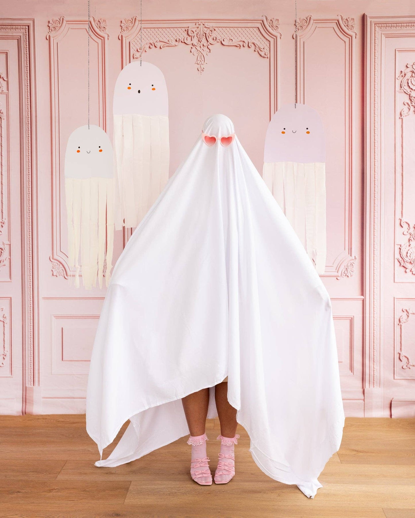 Person wearing a white ghost costume with pink shoes in front of a decorative pink wall with hanging ghost decorations.